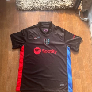 Svart FC Barcelona Nike fotbollströja - Snygg svart FC Barcelona fotbollströja från Nike med klubbmärke och röd Nike-logga på bröstet. Spotify-logga i rött framtill och röda samt blå sidopaneler. Tillverkad i lätt och ventilerande material, perfekt för match eller träning.