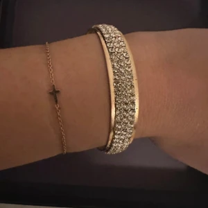 Glittrigt guldarmband med stenar - Snyggt armband i guldton med tre rader glittrande stenar. Armbandet är stelt och har en rund form