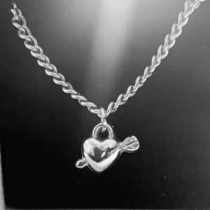 Silverhalsband med hjärthänge - Snyggt halsband i silverfärg med tvinnad kedja och ett hjärthänge som har en pil genom sig. Perfekt accessoar för att ge din outfit en romantisk touch. Passar dig som gillar stilrena smycken med en twist.