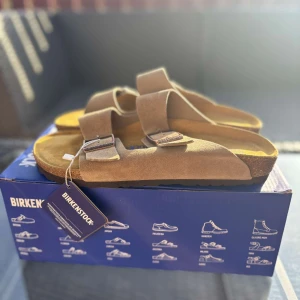 Birkenstock Arizona sandaler taupe mocka - Birkenstock Arizona sandaler i taupefärgad mocka med två breda remmar och justerbara metallspännen. Klassisk korksula och öppen tå för en chill och bekväm vibe. Perfekta för varma dagar och enkel att matcha med allt.
