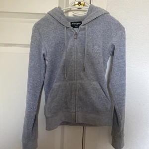 Grå hoodie från Juicy Couture XXS - Snygg grå hoodie från Juicy Couture i storlek XXS. Tröjan har huva, snörning och dragkedja framtill samt två fickor. Perfekt för en chill och avslappnad stil. Diskret Juicy Couture-logga broderad på bröstet.