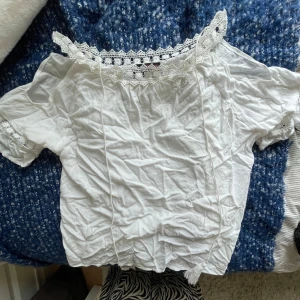 Vit blus med spets från EYN EYN - Superfin vit blus från EYN EYN med romantiska spetsdetaljer längs halsringningen och ärmsluten. Blusen har offshoulder-modell och korta ärmar, perfekt för en söt och feminin look. Tillverkad i mjuk bomull som känns lätt och luftig.