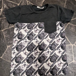 Svart t-shirt med hundmönster stl 128 - Cool svart t-shirt med korta ärmar och bröstficka. Nedre delen har ett unikt mönster med franska bulldoggar i svart och vitt. Perfekt för dig som älskar hundar och vill sticka ut med en rolig och annorlunda stil.