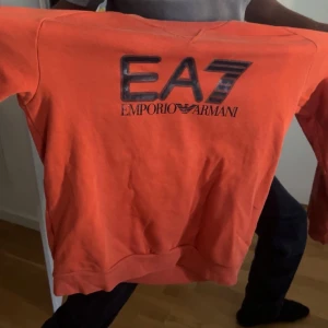 Orange EA7 Emporio Armani sweatshirt - Snygg orange sweatshirt från EA7 Emporio Armani med stor svart logga på bröstet. Tröjan har rund hals, ribbade muddar och är tillverkad i mjukt material som känns skönt mot huden. Perfekt för dig som vill sticka ut med en färgstark och trendig look.