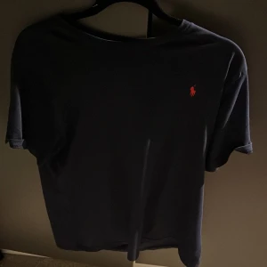 Mörkblå t-shirt från Polo Ralph Lauren - Snygg mörkblå t-shirt från Polo Ralph Lauren med klassisk röd broderad logga på bröstet. T-shirten är i mjuk bomull och har en normal passform med korta ärmar. Perfekt för en clean och stilren look. Det är en strl xl i barn men jag skulle säga att den passar dig som är mellan 178-185