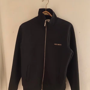Svart jacka från Les Deux med dragkedja - Svart jacka från Les Deux med stilren design och guldfärgad logga på bröstet. Jackan har hög krage, hel dragkedja framtill och två fickor. Tillverkad i mjukt material som känns skönt mot huden. Perfekt för dig som gillar en clean och sportig look.