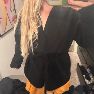 Svart stickad cardigan kofta med bälte - Mysig svart stickad cardigan jacka/kofta med bälte i midjan. Koftan har lång ärm och en avslappnad passform, perfekt att slänga på sig över en outfit för en chill och stilren look. Oanvänd