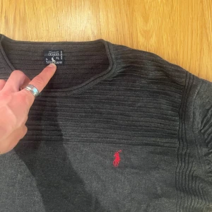 Mörkgrå stickad tröja Ralph Lauren - Mörkgrå stickad tröja från Ralph Lauren, passar large (L). Tröjan har ribbade partier på axlar och ärmar samt en röd broderad logga på bröstet. Klassisk rund halsringning och långärmad modell. Stilren.