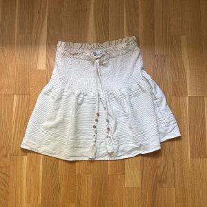 Vit kortkjol med snörning från Zara - Supersöt vit kortkjol från zaramed smockad midja och snörning med tofsar och pärlor framtill. Kjolen har volangkant och diskret struktur i tyget som ger en boho-vibe. Perfekt för varma dagar och enkel att styla med crop top eller linne. Jag köpte kjolen för något år sen när denna kjolen trendade men kände inte att jag använde den så mycket därför säljer jag den.💕💕