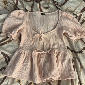 Ljusrosa blus med puffärm Gina Tricot - Supersöt ljusrosa blus från Gina Tricot med puffiga korta ärmar, v-ringning och två knytband framtill. Blusen har en flowy, volangig nederdel och är gjord i ett krinklat, mjukt tyg som känns luftigt och somrigt. Nästan aldrig använd och inga difekter. DM vid intresse💕