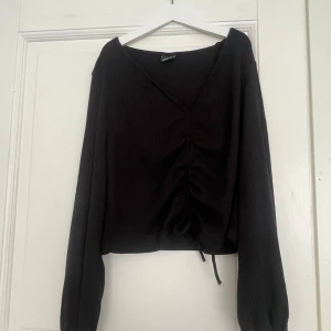 Svart ribbad tröja med snörning Gina Tricot - Svart långärmad tröja från Gina Tricot med ribbad struktur och v-ringning. Tröjan har snygg snörning framtill som ger en rynkad effekt och ballongärmar för en trendig look. Perfekt till jeans eller kjol för en chill stil.