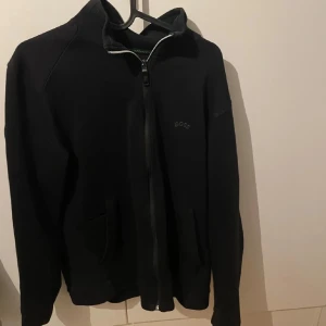 Svart zipjacka från BOSS - Svart jacka från BOSS med hel dragkedja framtill och hög krage. Jackan har diskret BOSS-logga på bröstet och två fickor. Perfekt för en clean och stilren look.