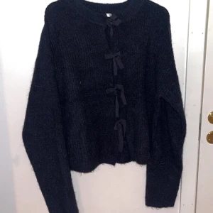 Svart stickad kofta med rosetter - Mysig svart stickad kofta med långa ärmar och fyra söta rosetter framtill. Koftan har en croppad passform och är perfekt för lager-på-lager-stil. Tjockt och fluffigt material som håller dig varm under kyliga dagar.