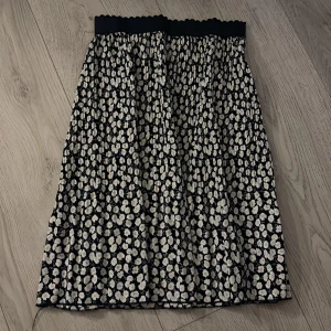 Blommig kortkjol från 2-Biz - Supersöt kortkjol från 2-Biz med svart botten och vita blommor. Kjolen har resår i midjan och är lätt rynkad för en skön och avslappnad look. 