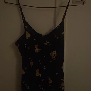 Svart långklänning med gula blommor - Supersöt svart långklänning med tunna axelband och blommigt mönster i gult och vitt. Klänningen har en slits framtill och är perfekt för varma dagar. Materialet känns lätt och flowy, och modellen är figurnära upptill med lösare fall nedtill.