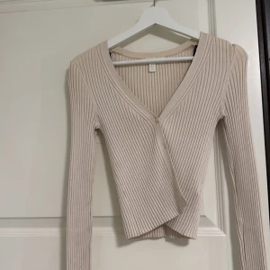 Beige ribbad omlottkofta från H&M - Säljer en beige ribbad omlottkofta från H&M i storlek XS. Koftan har v-ringning, lång ärm och stängs med en knapp framtill. Den är croppad och har en snygg, figurnära passform som passar perfekt till jeans eller kjol.