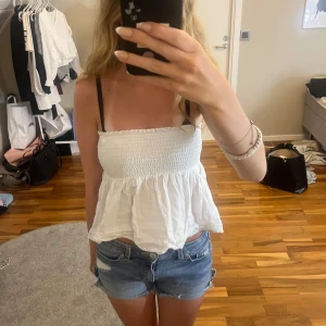 Vit bandeau tubtopp från Gina Tricot - Supersöt vit bandeau tubtopp från Gina Tricot med smockad överdel och volangig nederkant. Toppen är ärmlös och har en luftig, flowy passform som är perfekt för varma dagar. Enkel att matcha med shorts eller kjol för en chill sommarlook. Skrynklig men det går lätt att stryka den. Skulle finnas band till men de är tyvärr borta.