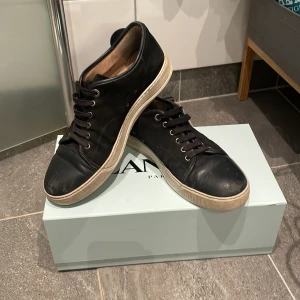 Svarta sneakers från Lanvin i skinn - Stilrena svarta sneakers från Lanvin i skinn med mockadetaljer och vita gummisulor. Storlek 43 men passar även 44. Klassisk rund tå, svarta skosnören och diskret logga på sulan. Kommer med originalkartong, extra skosnören och dustbag. Perfekta för dig som gillar lyxiga och tidlösa sneakers. Skick 6.5/10, pris går att diskutera.
