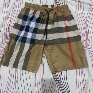 Rutig badshorts från Burberry - Snygga badshorts från Burberry med klassiskt rutigt mönster i beige, brunt, svart, vitt och rött. Shortsen har elastisk midja med svart snörning och är perfekta för sommaren. Ikonisk design som sticker ut på stranden.