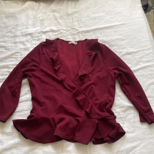 Vinröd omlottopp med volang - Supersnygg vinröd omlottopp från Nelly med volangdetaljer längs ringningen och nederkanten. Topp med trekvartsärm och knäppning i sidan. Passar jätte bra med en turtleneck under 