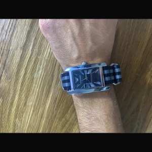 Emporio Armani rektangulär klocka - Stilren klocka från Emporio Armani med rektangulärt silverfärgat boett och svart urtavla med romerska siffror. Klockan har ett randigt svart och grått tygarmband med metallspännen. Perfekt accessoar för dig som gillar klassisk design med modern twist.