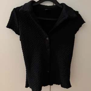 Svart kortärmad blus med knappar - Svart blus med korta ärmar och krage. Toppen har en strukturerad, lite bubblig yta och knäpps framtill med svarta knappar. Perfekt till jeans eller kjol för en stilren look.