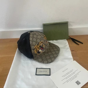 Gucci keps med tiger och GG-mönster - Snygg keps från Gucci med klassiskt GG-monogram i beige och brunt, broderade tigrar framtill och svarta mesh-paneler på sidorna. Märkeslapp på insidan och böjd skärm. Perfekt statement-piece för dig som vill sticka ut.