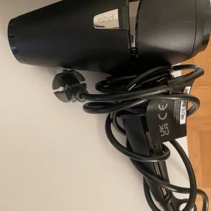 Svart hårfön från GHD - Snygg svart hårfön från GHD med stilren design och blank detalj. Kommer med lång sladd och brittisk kontakt. Perfekt för att snabbt styla håret och få till en proffsig look hemma. Använd typ en gång då jag fick en annan typ av hårfön av en vän