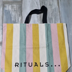 Randig shoppingväska från Rituals - Färgglad shoppingväska från Rituals med breda ränder i gult, rosa, grönblått och vitt. Väskan har svarta handtag och tryck på båda sidor, ena med texten 'RITUALS...' och andra med 'SMILE AND THE WORLD SMILES BACK'. Tillverkad i kraftig bomull.