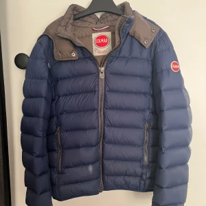 Blå dunjacka från Colmar, 154 cm - Snygg blå dunjacka från Colmar med bruna detaljer vid kragen. Jackan har quiltad design, dragkedja framtill och två fickor med dragkedja. Perfekt för kalla dagar och har en sportig vibe. Klassisk logga på bröstet och ärmen. Har två slitningar (se bild) som är lagade med hjälp av genomskinliga klisterlappar.