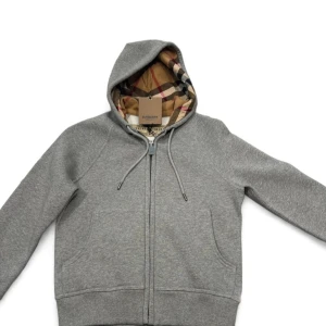 Grå burberry hoodie - Snygg grå hoodie från Burberry med klassiskt rutigt foder i huvan. Tröjan har hel dragkedja, snörning i huvan och två fickor framtill. Perfekt för dig som gillar stilrena och exklusiva plagg med en twist.