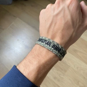 Christian Dior J'Adior armband - Snyggt armband från Christian Dior med texten 'J'Adior' broderad i svart. Armbandet har camouflage-mönster i beige, grönt och brunt och är gjort i ett vävt tygmaterial med fransar i ändarna. Perfekt accessoar för att lyfta din outfit.