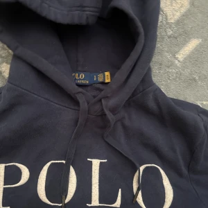 Mörkblå hoodie från Polo Ralph Lauren - Snygg mörkblå hoodie från Polo Ralph Lauren med stor vit logga framtill och klassisk huva med snörning. Tröjan har känguruficka och långa ärmar, perfekt för en chill och stilren look. Materialet är mjuk bomull som känns skönt mot huden.