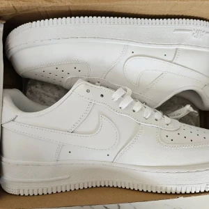 Nike Air Force 1 - Klassiska Nike Air Force 1 sneakers i helvitt skinn. Snygg och clean design med perforerad tå, vit sula och ikonisk swoosh på sidorna. Lågt skaft och snörning framtill. Perfekta för dig som vill ha en tidlös och fräsch look. En del smuts på skorna med det går bort om du tvättar i nån minut! 