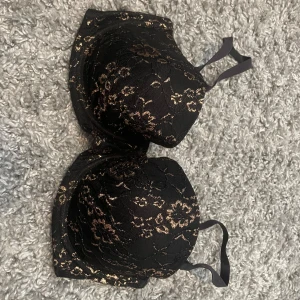 Svart och guldig bandeau tubtopp - Säljer en snygg bandeau tubtopp i svart med guldiga blommiga detaljer. Toppen har avtagbara axelband och är täckt av spets med glittriga inslag. Perfekt för dig som vill sticka ut lite extra med en elegant och trendig look.