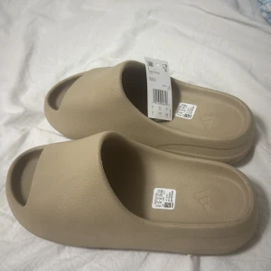 Yeezy slides pris kan diskuteras  - Säljer ett par beige adidas Yeezy Slide sandaler med chunky, vågformad sula och öppen tå. De är tillverkade i syntetmaterial och har en minimalistisk design med adidas-logga på fotbädden. Perfekta för chill dagar eller när du vill ha en clean look.