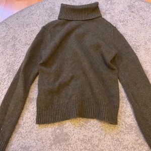 Mörkgrön stickad polotröja - Mysig mörkgrön stickad polotröja med hög krage och långa ärmar. Tröjan har ribbade muddar vid ärmslut och nederkant, perfekt för kyliga dagar. Enkel och stilren design som är lätt att matcha med olika outfits.