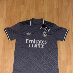 Real Madrid x Adidas grå fotbollströja M - Snygg grå Real Madrid fotbollströja från Adidas med diskret mönster, klassisk krage och vita detaljer på ärmslut och krage. Broderad klubbmärke och Adidas-logga på bröstet samt 'Emirates Fly Better' tryck på framsidan. Tillverkad i lätt och ventilerande polyester.
