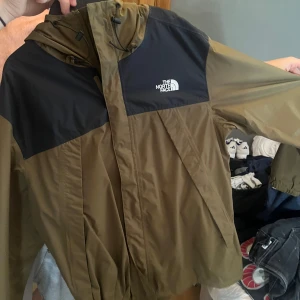 The North Face vindjacka brun/svart - Snygg vindjacka från The North Face i brunt och svart. Jackan har huva med snörning, dragkedja framtill och logga på både bröst och rygg. Perfekt för blåsiga dagar och enkel att matcha med streetwear. Lätt och smidig att ta med sig.