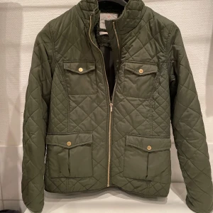 Olivgrön quiltad jacka Holly & Whyte - Snygg olivgrön quiltad jacka från Holly & Whyte by Lindex. Jackan har fyra fickor med lock och guldiga knappar, dragkedja framtill och mjukt foder. Perfekt för dig som vill ha en stilren och bekväm jacka med coola detaljer.