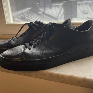 Svarta sneakers från Axel Arigato - Snygga svarta sneakers från Axel Arigato i croco-präglat skinn. Skorna har rund tå, platt sula och svarta snören. De är lite slitna på insidorna längst in men det är inget man tänker på. Också lite gångväck och slitet där fram som jag fotade