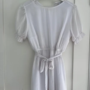 Vit klänning med puffärm och knytband i midjan - Vit klänning från NA-KD med puffärm och knytband i midjan. Det finns en fläck på ryggen - syns inte så bra på bild, även något liknande på bandet i midjan som ser ut som fett fläckar men har ingen aning. Jag själv har aldrig använd klänningen men den är begagnad. Klänningen är lite genomskinlig men annars i ändå helt okej/bra skick