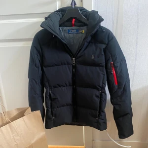 Polo Ralph Lauren Dunjacka - Snygg svart dunjacka från Polo Ralph Lauren i storlek XS. Jackan har en klassisk pufferstil med huva, dragkedja framtill och en röd detalj på ärmfickan. Broderad logga på bröstet och flera praktiska fickor. Perfekt för kalla dagar och riktigt stilren look. Nypris: 5495kr. Marinblå/svart. OBS - priset är högt på grund av plicks nya system med deras avgift de tar. Billigare på vinted.