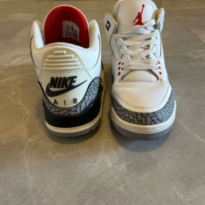 Nike Air Jordan 3 Retro White Cement - Nike Air Jordan 3 Retro White Cement med vit ovandel i skinn, ikoniska grå elefantmönstrade paneler och svarta detaljer. Röda accenter på snörhål och Jumpman-logga. Grå yttersula och röd insida ger en klassisk look. Perfekta för dig som gillar streetwear och sneakerskultur. Skorna har lite repor be om fler bilder ifall du vill se de bättre. 
