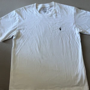 Vit t-shirt från Polo Ralph Lauren - Klassisk vit t-shirt från Polo Ralph Lauren med korta ärmar och rund hals. Ikoniska lilla broderade loggan på bröstet. Skön och mjuk bomull, perfekt till jeans eller shorts. Enkel och stilren vibe.