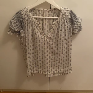 Vit blommig blus med volangärm - Luftig vit blus med småblommigt mönster i mörkblått och volangiga korta ärmar. V-ringad hals och extra detaljer på ärmarna med paisleyinspirerat tryck. Perfekt för en bohemisk vibe och sköna sommardagar.