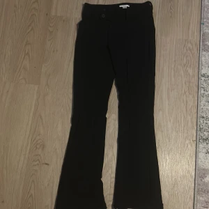 Svarta bootcut kostymbyxor - Säljer ett par svarta kostymbyxor med bootcut-snitt och hög midja. Byxorna har två knappar i midjan och bälteshällor. Snygga och stilrena, perfekta till en trendig look.