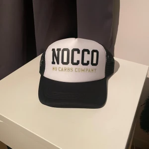 Svartvit NOCCO keps med mesh - Snygg svart och vit keps från NOCCO med tydlig logga framtill och texten 'NO CARBS COMPANY'. Kepsen har klassisk trucker-stil med svart böjd skärm och svart mesh på sidorna och bak, vilket ger bra ventilation. Perfekt för dig som gillar sportig och trendig stil.  Helt ny skick limiterad aldrig använd 