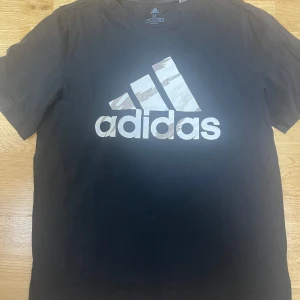 Svart Adidas t-shirt med logga - Svart t-shirt från Adidas med stor vit logga framtill. Klassisk rund halsringning och korta ärmar. Tillverkad i mjuk bomull, perfekt för en sportig och avslappnad stil. Passar dig som gillar streetwear och sportmode.
