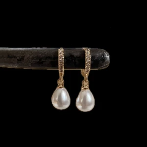 Guldiga örhängen med droppformade pärlor - Eleganta örhängen i guldton med små gnistrande stenar på ringen och droppformade vita pärlor som hänger ned. Perfekta för att lyfta din stil med en klassisk och lyxig touch.